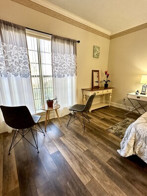 A room with a bed, two chairs, a small table with a plant, a desk with a lamp, and a window with curtains.