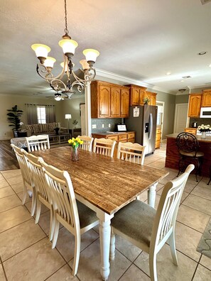 A spacious dining area with a wooden table and white chairs, a kitchen with wooden cabinets, and a chandelier.
