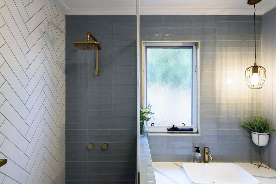 Un baño moderno con un patrón de baldosas en espiga, ducha con grifo dorado, ventana con vistas, lavamanos blanco y una lámpara colgante.