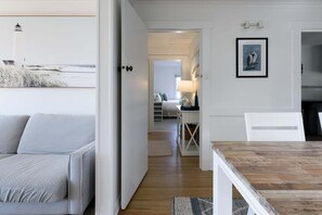 Een kamer met een houten eettafel, een witte bank, een lijst met een foto aan de muur, en uitzicht op een strand met een vuurtoren.