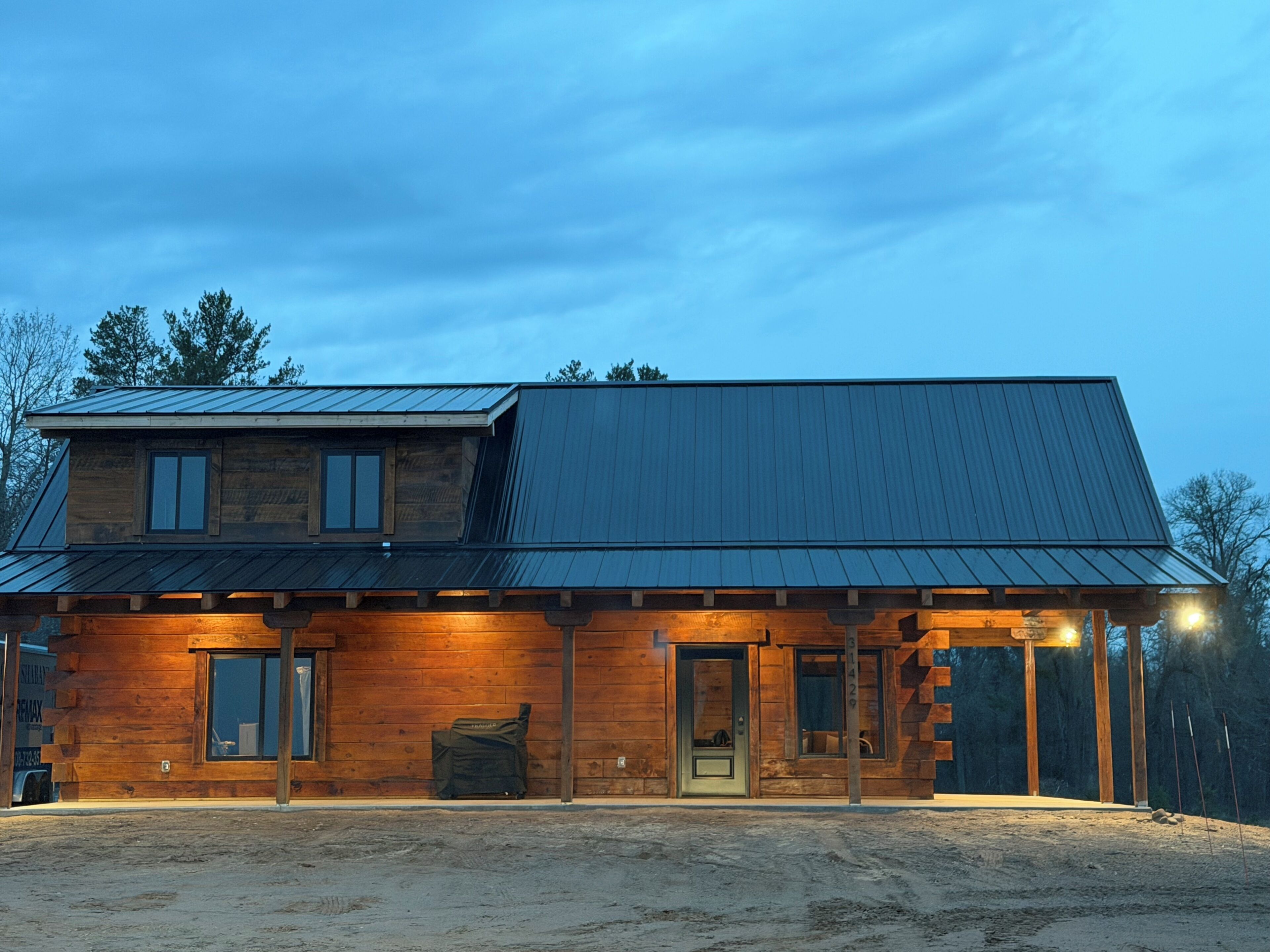 A two-story wooden cabin with a metal roof, a covered porch, and a dirt ground.