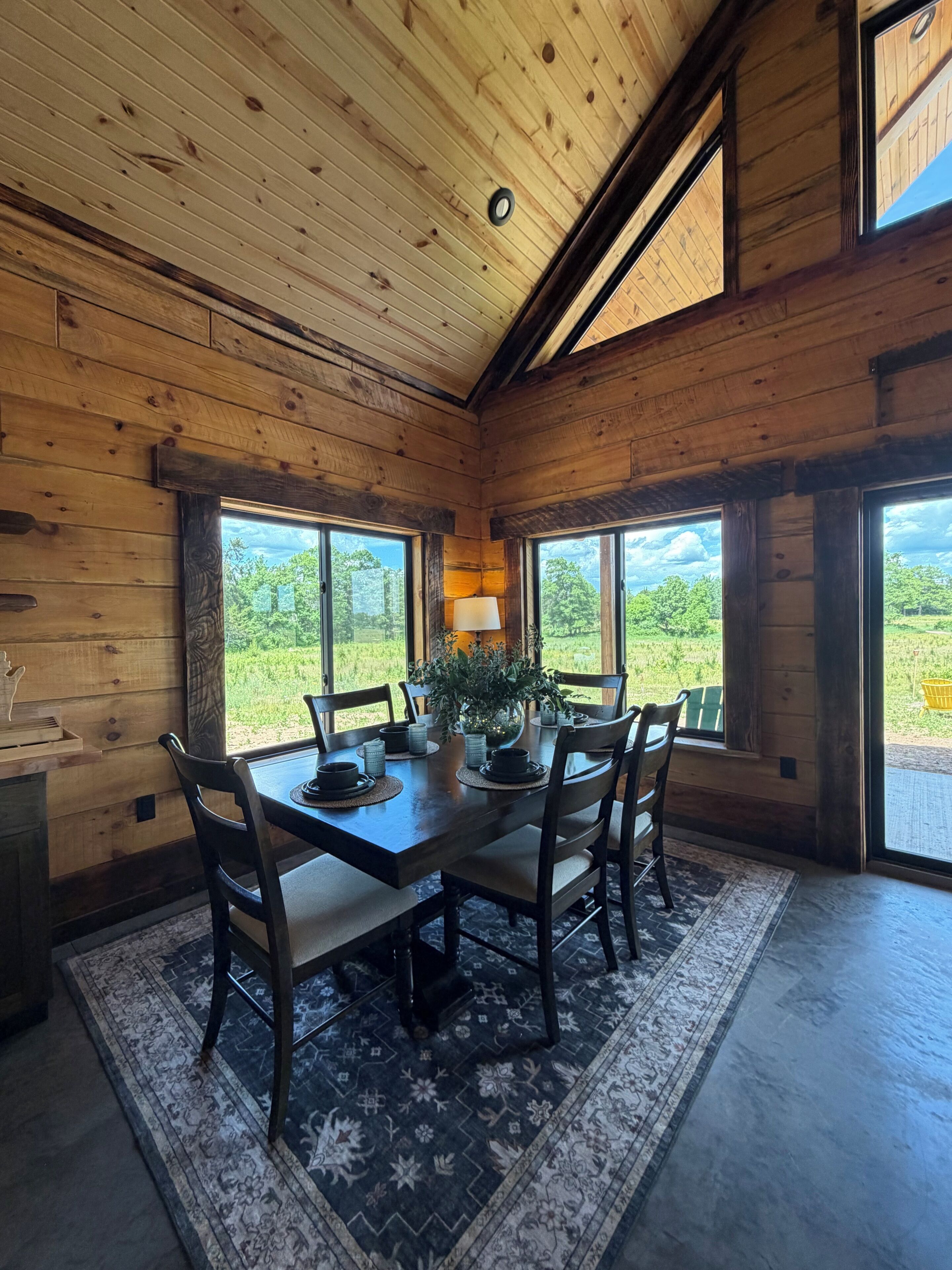 Un interior de madera con una mesa de comedor dispuesta para seis personas, ventanas amplias y vistas al exterior con vegetación.