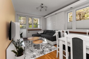 A modern living room with a sectional sofa, a dining table with chairs, a TV mounted on the wall, and large windows with blinds. - Leśny Zakątek by Grand Apartments (Sopot)