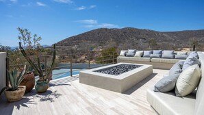 A rooftop terrace with a stone fire pit, a sofa with cushions, potted plants, and a view of a mountainous landscape. - Modern 4BR Villa with Rooftop & Pickleball (Los Barriles)