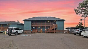 A building with a ramp and a staircase leading to a balcony, a parking lot with several cars, and a sunset sky. - Sunset Haven (Depoe Bay)