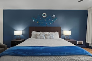Una cama bien tendida con una manta azul, dos lámparas de noche, un adorno en la pared y una mesita de noche con una lámpara.