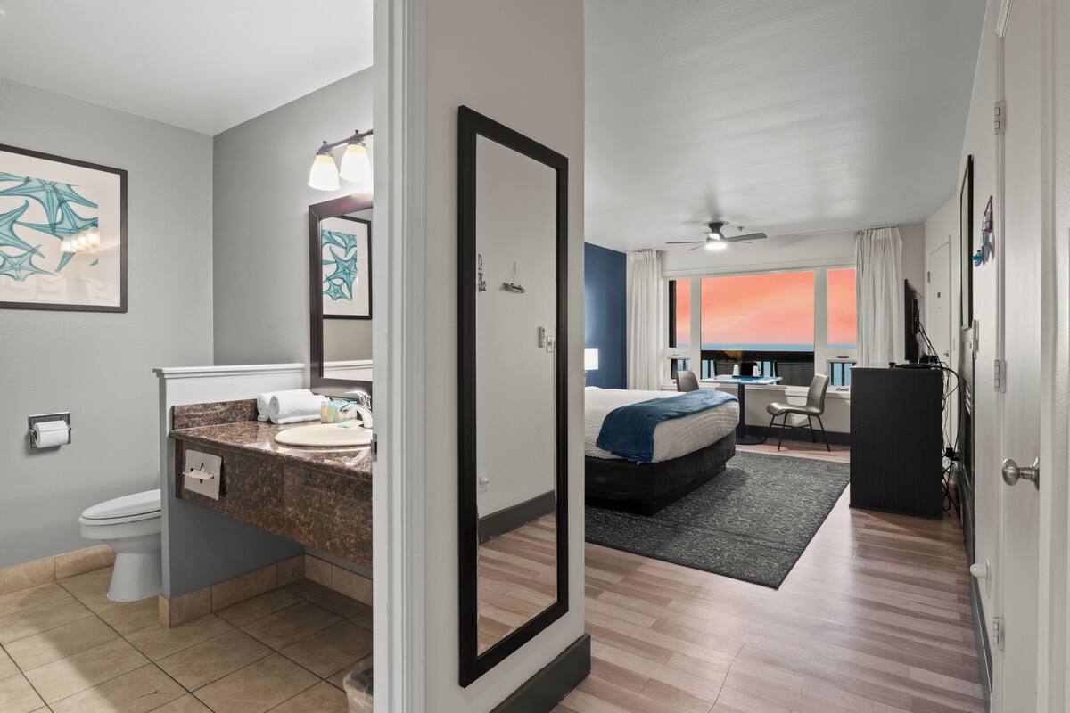 A modern hotel room with a bathroom, a bed, a desk, and a view of the sunset.