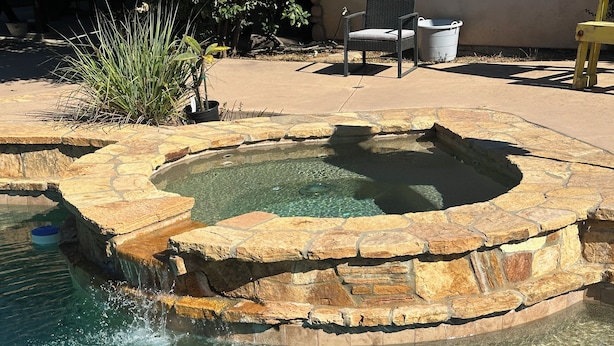 A stone hot tub with clear water, surrounded by a stone deck, and a chair is visible in the background.