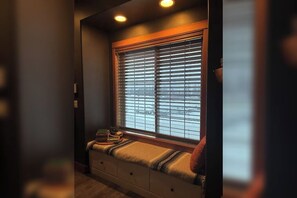 Ein Fenster mit Jalousien, eine Bank mit Decke und ein kleiner Tisch mit Büchern.