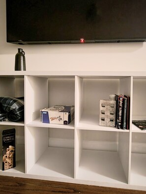 Ein weißes Regal mit einem Flachbildfernseher darüber, einer Metall-Wasserflasche, einer Taschentuchbox, einem Buch mit dem Titel 'Unfinished Glenjones' und einer kleinen Figur.