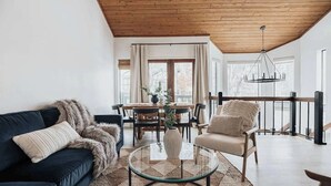 A modern living room with a dark blue sofa, a glass coffee table, a beige armchair, and a dining area with a wooden table and chairs. - Bearfoot Lake Escape (Big Bear Lake)
