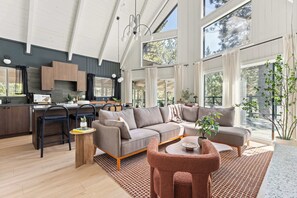 A spacious living area with a large sofa, a coffee table, and a dining table. There is a kitchen area with modern appliances and a large window providing natural light. - A-Frame in Moonridge, Big Bear Lake - A/C, Hot Tub (Big Bear Lake)