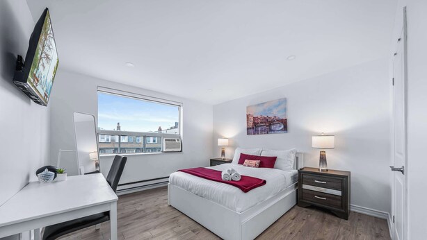 Une chambre moderne avec une grande fenêtre, un bureau, un lit avec un plaid rouge et une table de chevet avec une lampe.