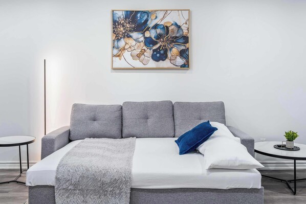 A modern bedroom with a grey sofa bed, a white bedspread, a blue pillow, a small round table with a plant, and a wall-mounted abstract painting.