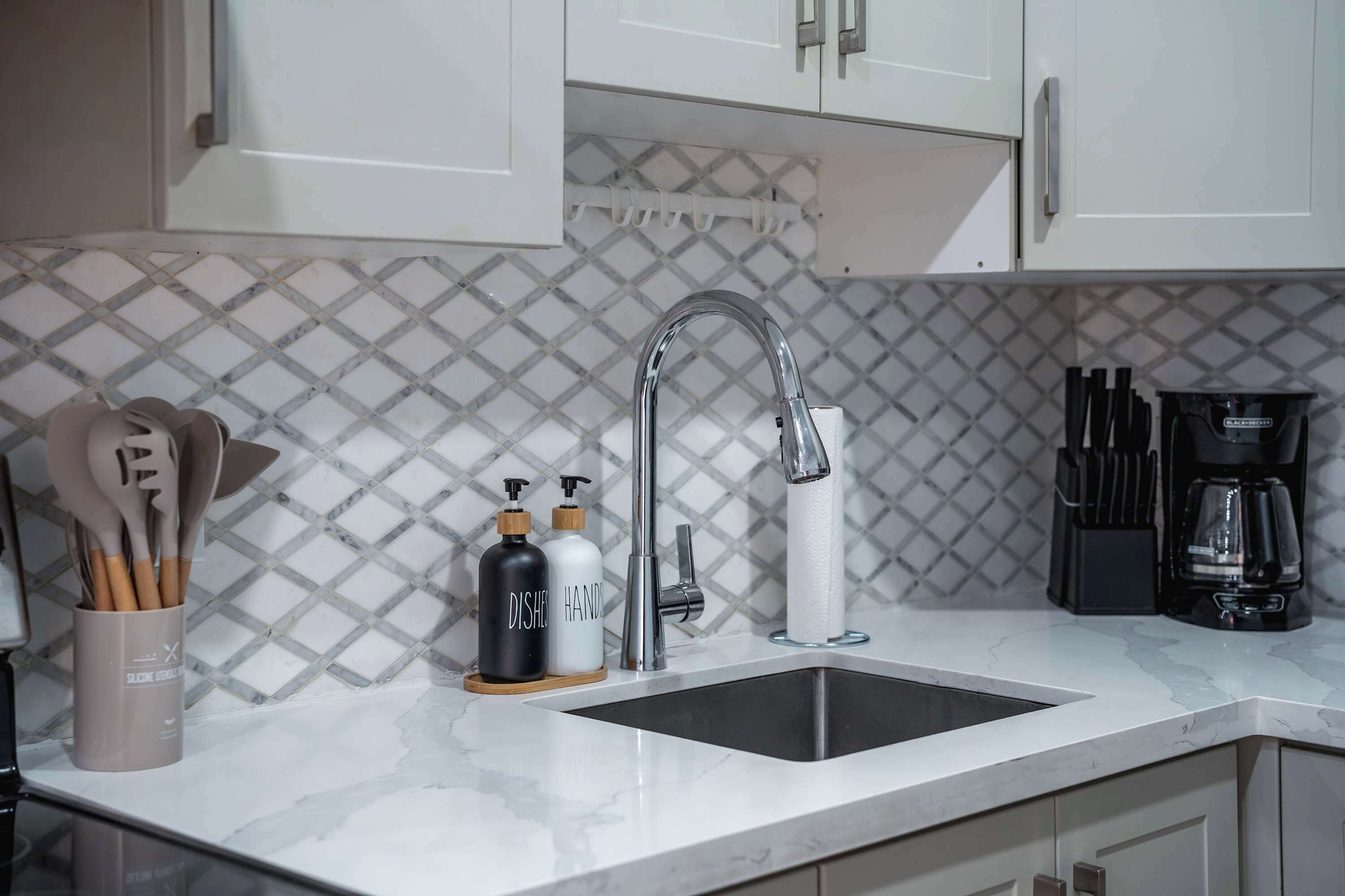 A kitchen with a sink, white cabinets, a patterned backsplash, and various kitchen utensils and appliances.
