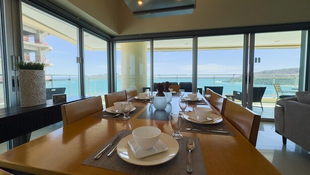 Uma área de refeições com uma mesa posta para quatro pessoas, grandes janelas com vista para o mar e um vaso de planta no parapeito.