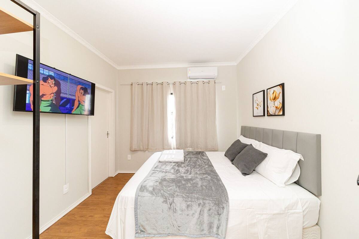 Quarto com cama, televisão, janela com cortinas e ar-condicionado fixado na parede.