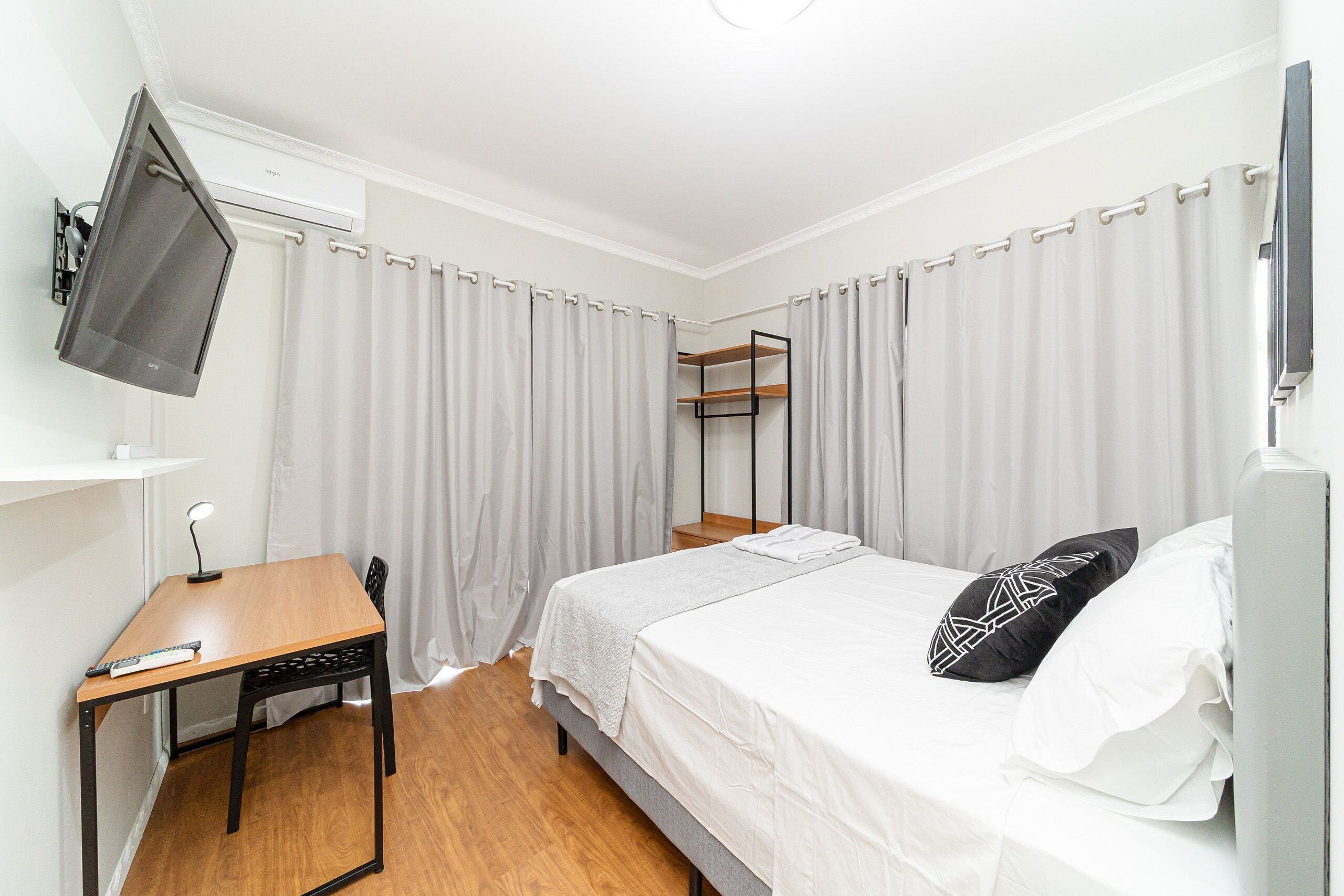 A bedroom with a bed, a desk, a TV mounted on the wall, and a shelf unit.