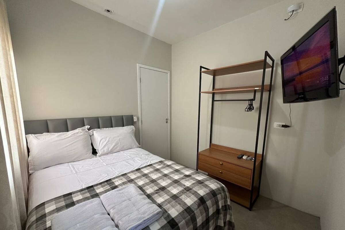 Quarto com uma cama, mesa de cabeceira, TV fixada na parede e uma porta.