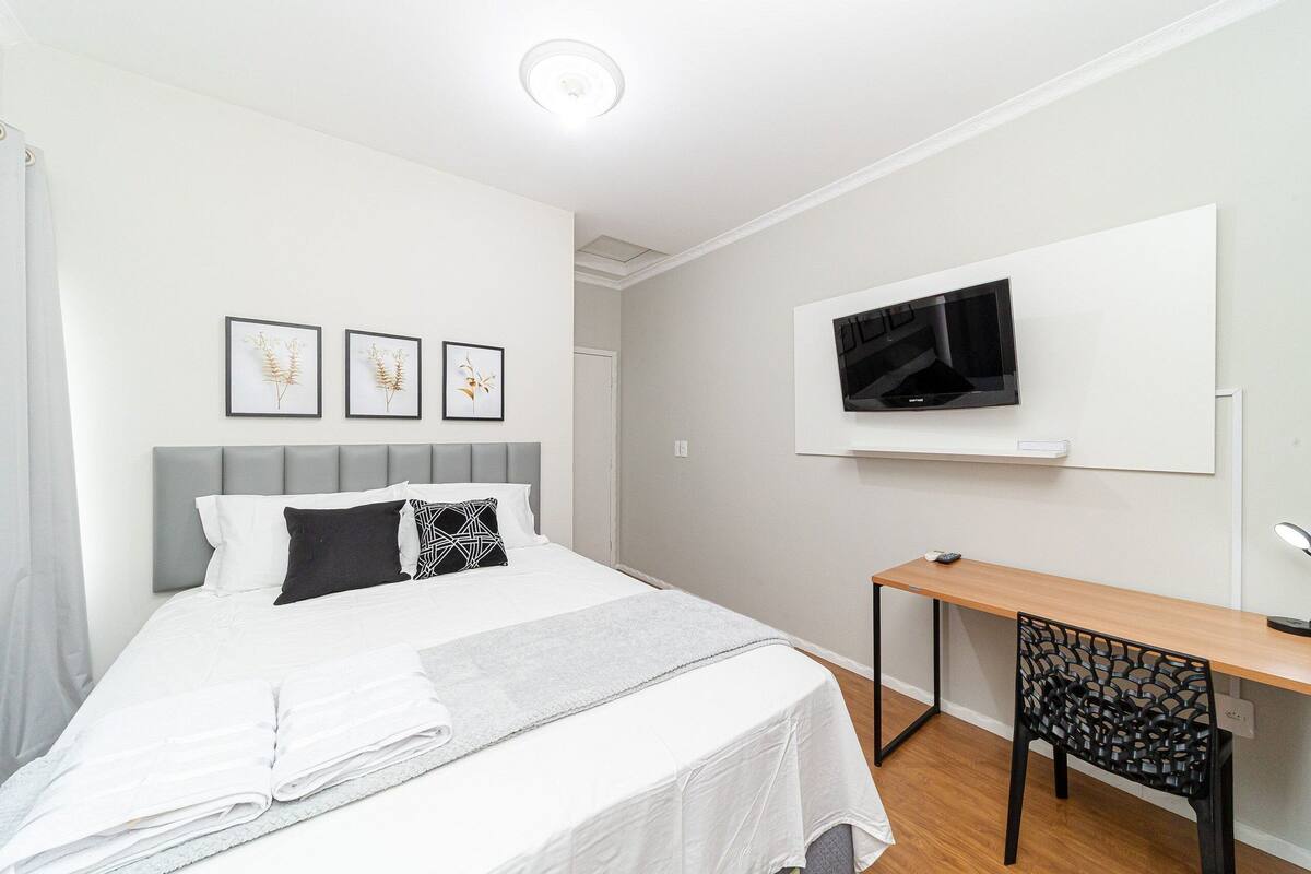 Quarto com cama, escrivaninha, cadeira, televisão e quadros nas paredes.