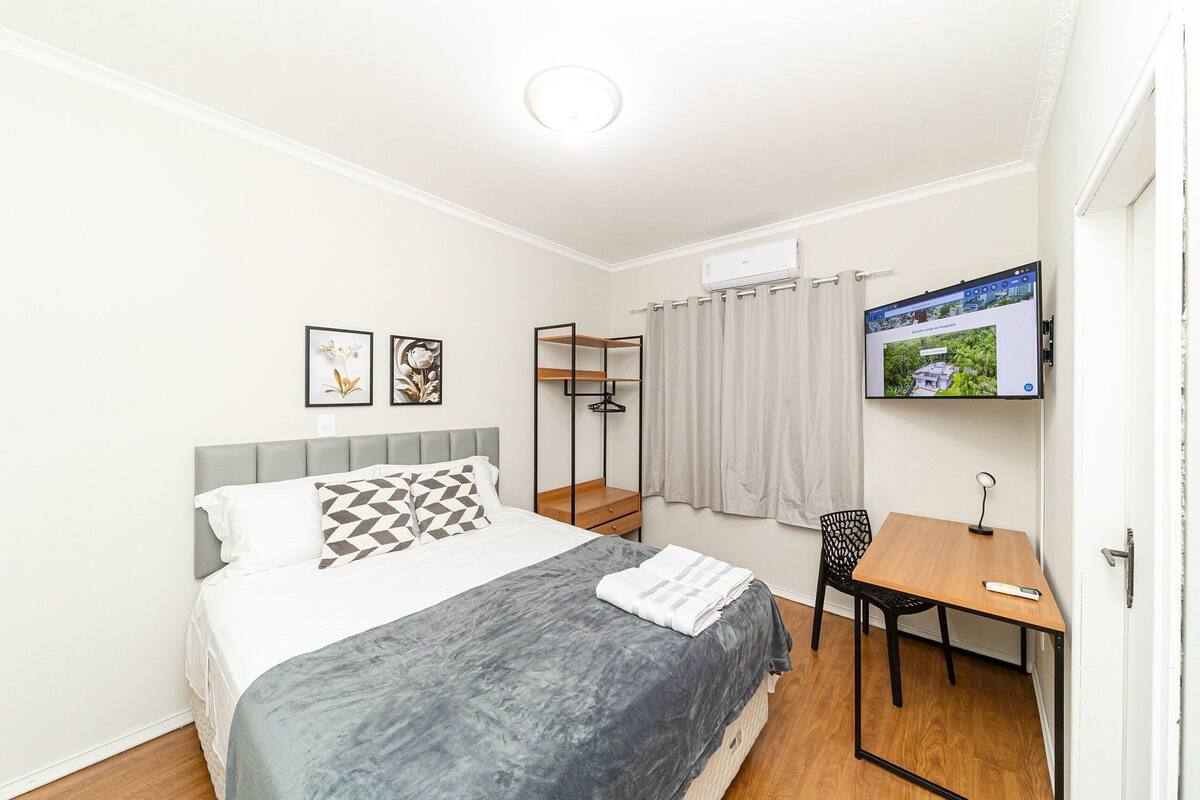 Quarto com cama, escrivaninha, cadeira, televisão e janela com cortinas.