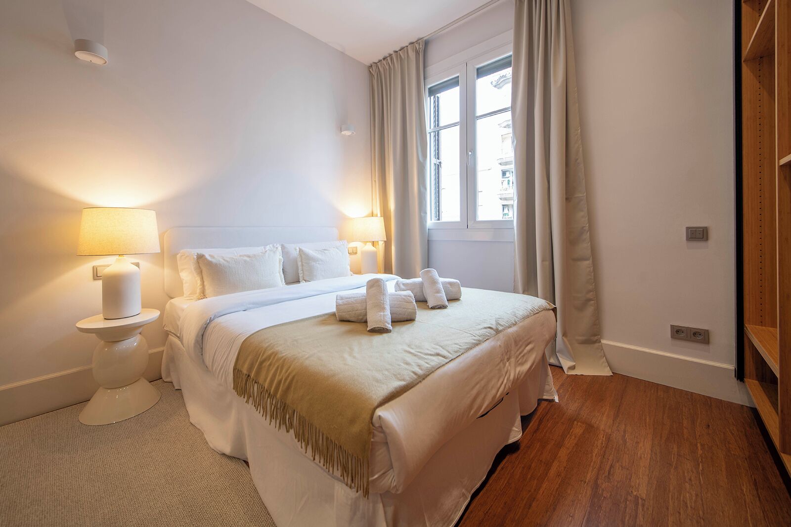 Ein Hotelzimmer mit Bett, Nachttischlampen, einem Fenster mit Vorhängen und Holzboden.