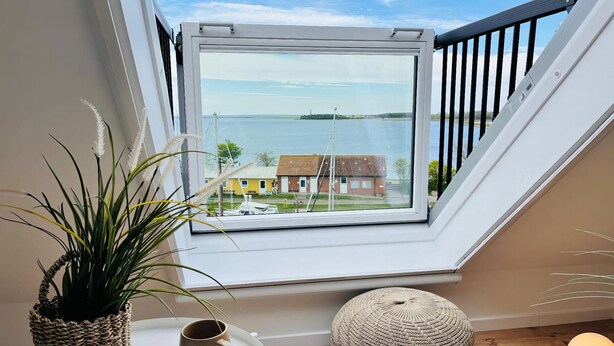 A room with a view of a body of water, a window with a black railing, a round woven ottoman, and a small table with a plant and a cup. - Ferienwohnung in Orth auf Fehmarn (Fehmarn)