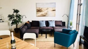 A modern living room with a sectional sofa, a blue armchair, a wooden dining table with a bottle and glasses, a small round table, and a large window with curtains. - Ferienwohnung in Orth auf Fehmarn (Fehmarn)