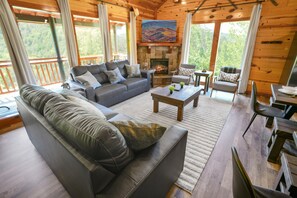 A cozy cabin interior with a fireplace, a sofa, a dining table, and a view of the forest. - Luxury Cabin, Sleeps 22 w/ Pool, Game Room, Movie Theater! (Sevierville)