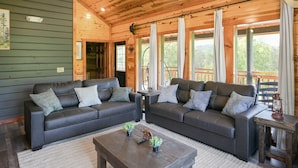 Een houten cabin met daarin twee donkerbruine leren banken, een houten salontafel en uitzicht op een houten terras door grote ramen.