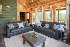 A wooden cabin interior with two dark leather sofas, a wooden coffee table, and a view of a wooden deck through large windows. - Luxury Cabin, Sleeps 22 w/ Pool, Game Room, Movie Theater! (Sevierville)