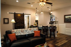 A living room with a black leather sofa, wooden chairs, a chandelier, and a kitchen area with a white countertop and blue dishware.