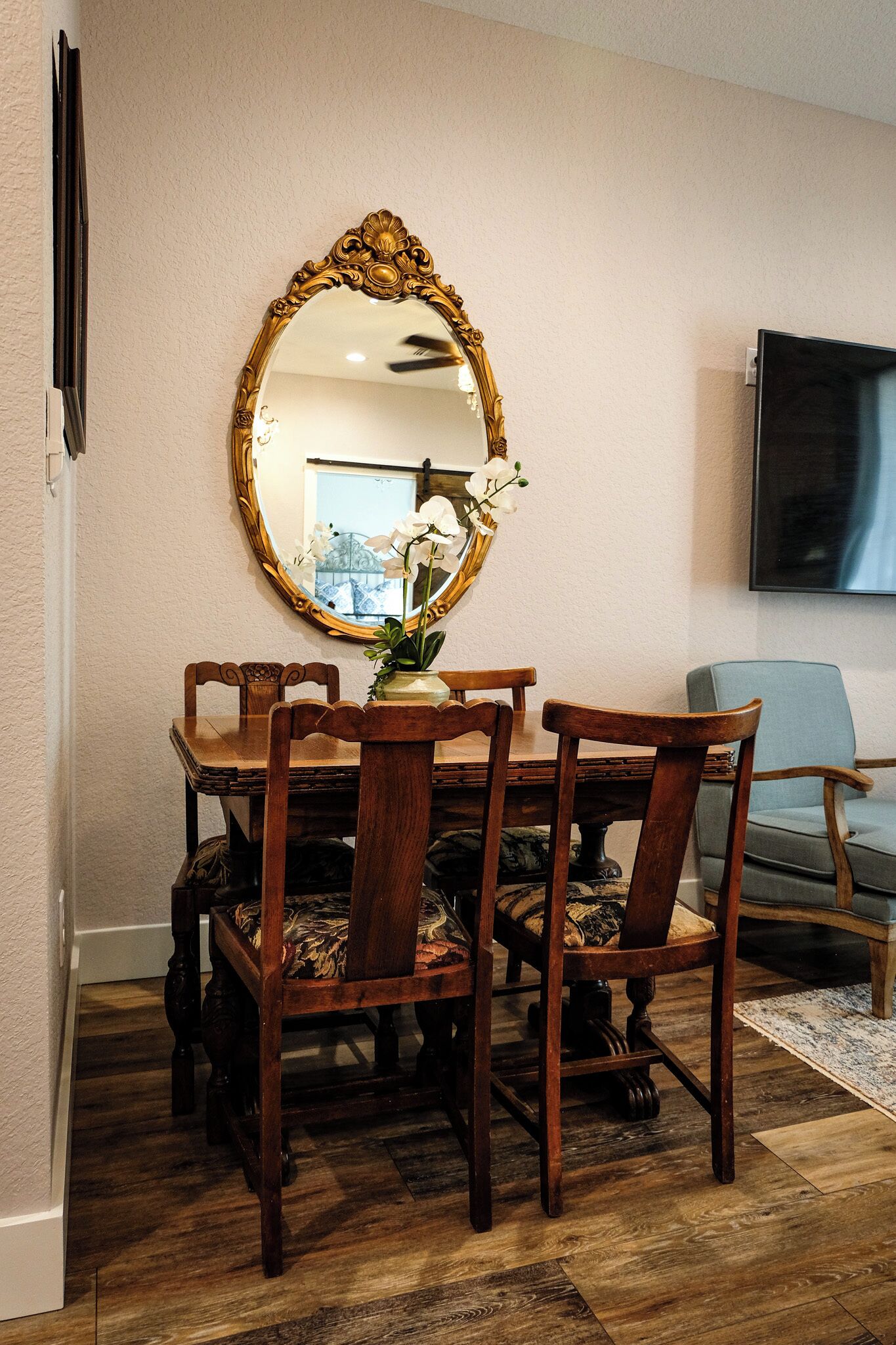 A dining area with a wooden table surrounded by chairs, a large ornate mirror on the wall, and a television mounted above.
