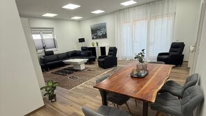 Uma sala de estar moderna com uma mesa de jantar de madeira, cadeiras estofadas em cinza, um sofá de couro preto e uma mesa de centro branca.