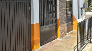 Uma entrada murada com portão de metal e porta, um caminho de concreto e um edifício com parede de tijolos e janela.