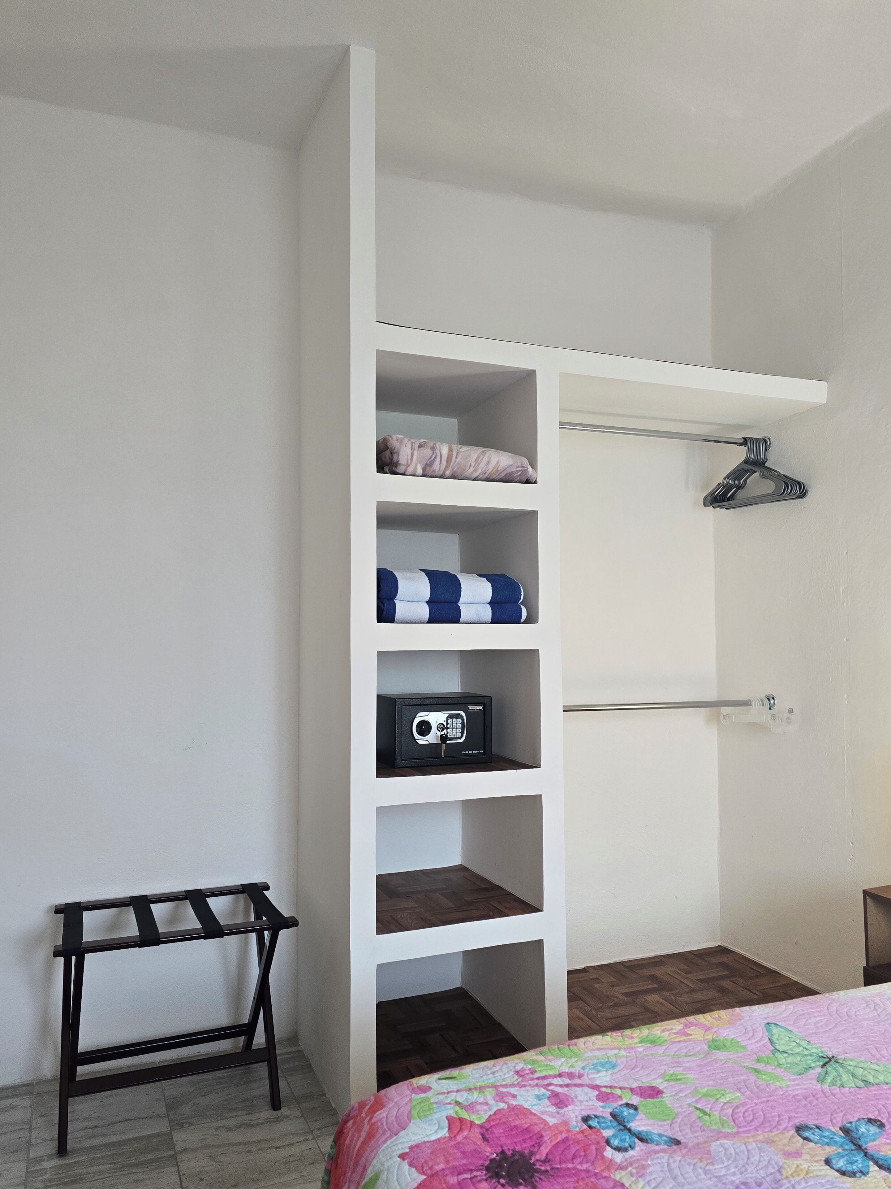 Un dormitorio con cama, armario con varios estantes, barra para colgar ropa y una mesita.