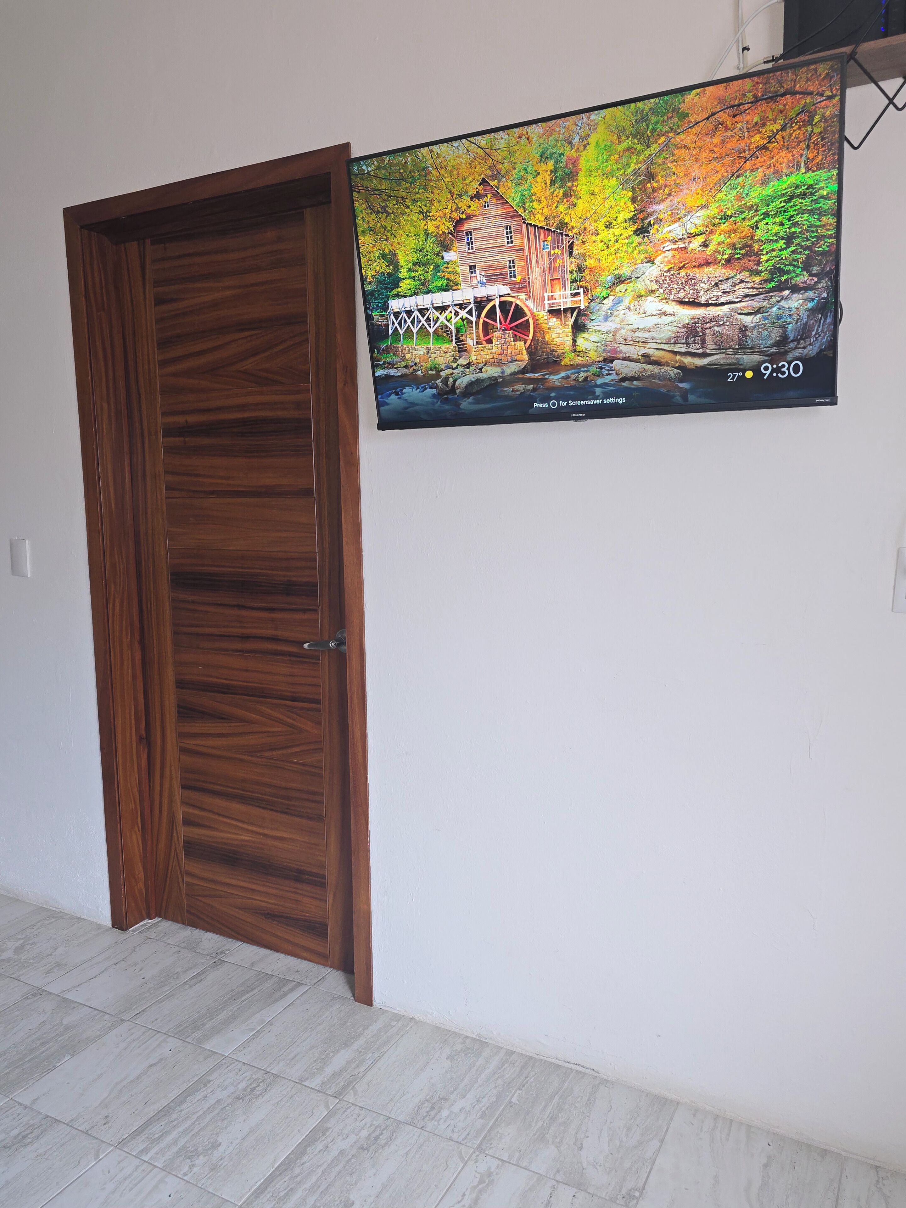 A room with a wooden door, a tiled floor, and a wall-mounted television displaying a scenic image.