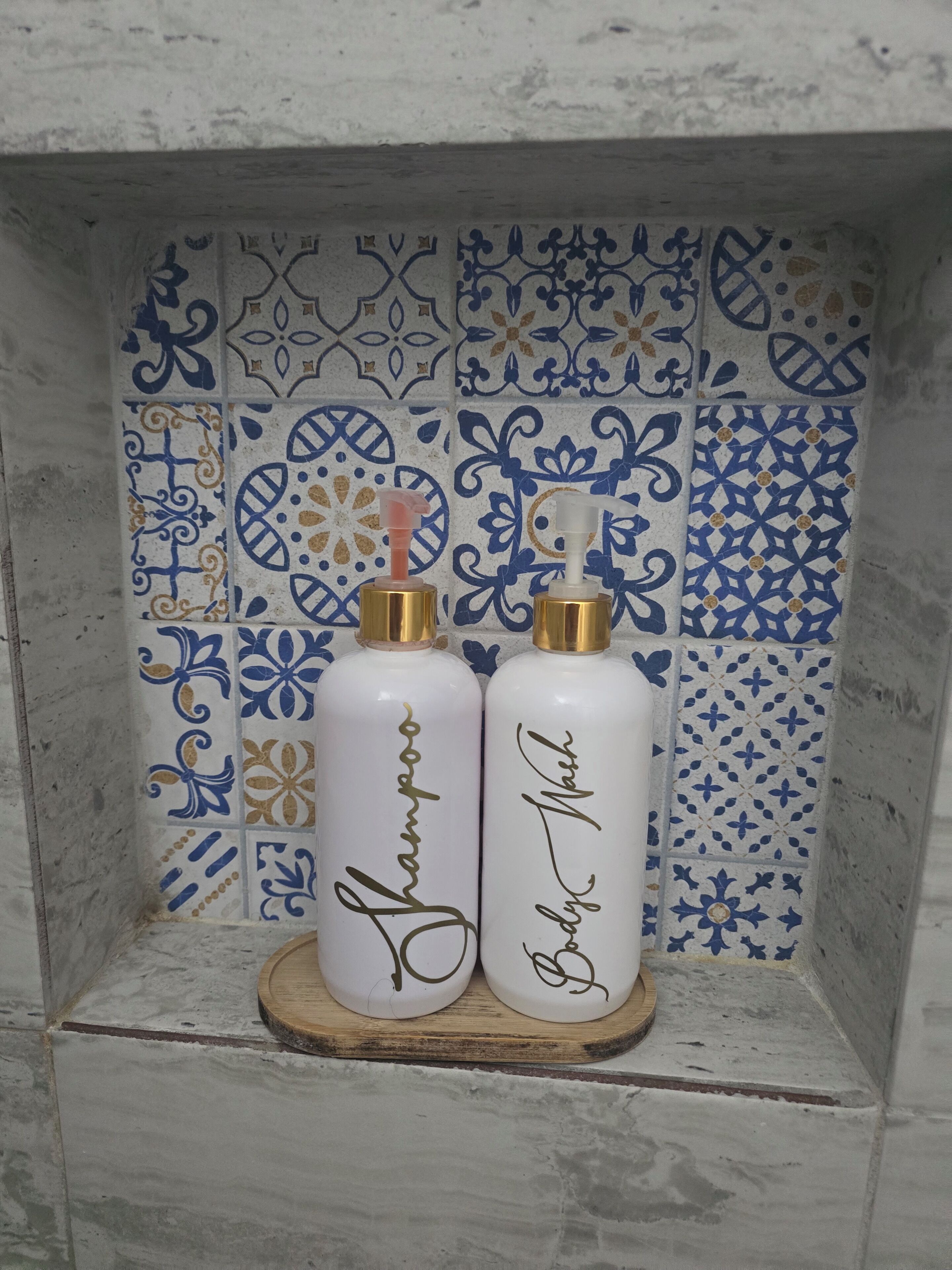 Two white bottles with gold caps labeled 'Shampoo' and 'Body Wash' on a wooden tray against a tiled wall with blue and white patterns.