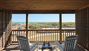A wooden deck with two chairs and a table, overlooking a beach and a wooden structure. - Ocean View King + Restaurants & Pool + Spa (Duck)