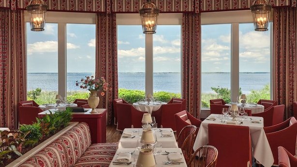 Un ristorante con tavoli apparecchiati, ampie finestre vista mare e tende a motivi decorativi.