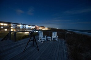 Een telescoop op een houten terras met stoelen, met uitzicht op een strand en gebouwen.