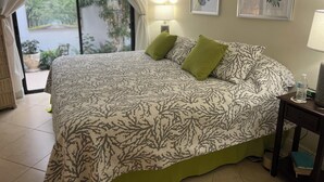 Un lit avec une couvre-lit orné de motifs de feuilles, un oreiller vert, une table de chevet avec une lampe et, au mur, des œuvres d’art encadrées représentant des feuilles.