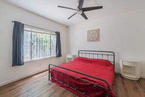 Ein Schlafzimmer mit Bett, Deckenventilator, Fenster mit Vorhängen, Nachttisch und Wanddeko.