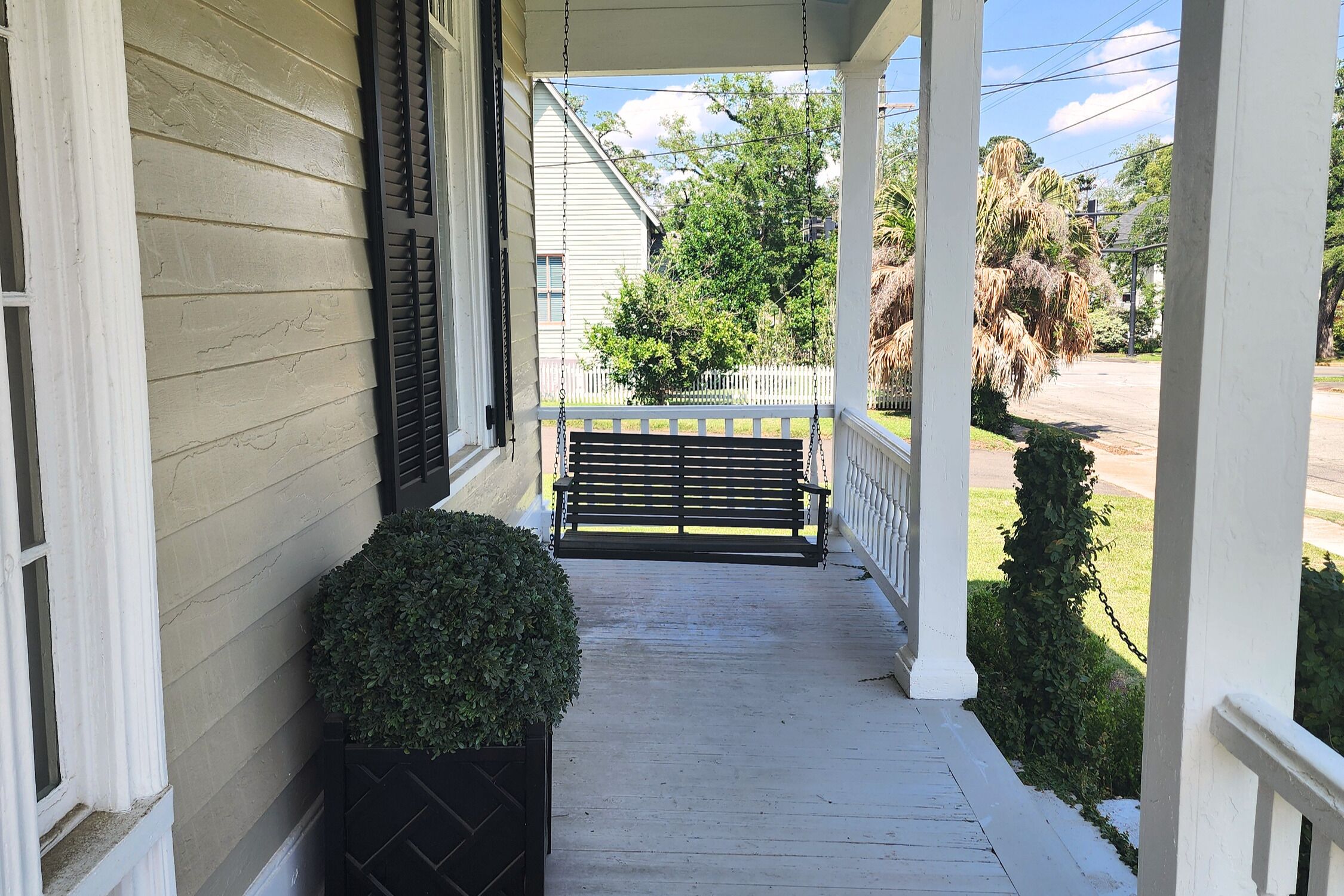 A porch with a bench, a potted plant, and a view of a white fence and greenery.