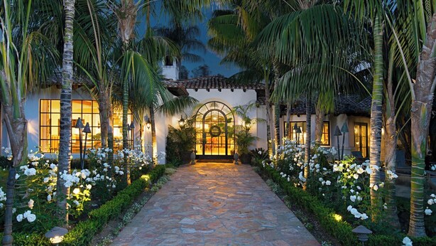 A pathway leading to a building with a tiled roof and a central archway, surrounded by palm trees and white flowers. - Gated 7BR Rancho Santa Fe Estate Pool/Guest House (Rancho Santa Fe)
