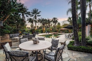 An outdoor patio with a round table and chairs, surrounded by palm trees and a pool.