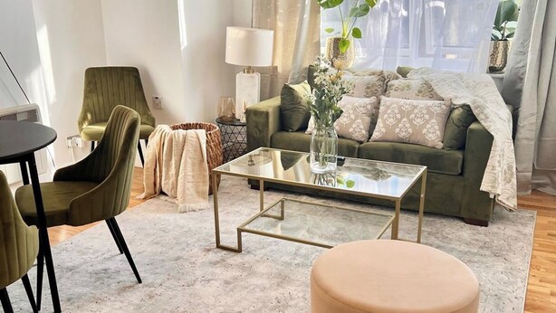 Ein Wohnzimmer mit einer Couch, einem Couchtisch, einem runden Hocker, einer Lampe und Blick ins Freie durch transparente Vorhänge.
