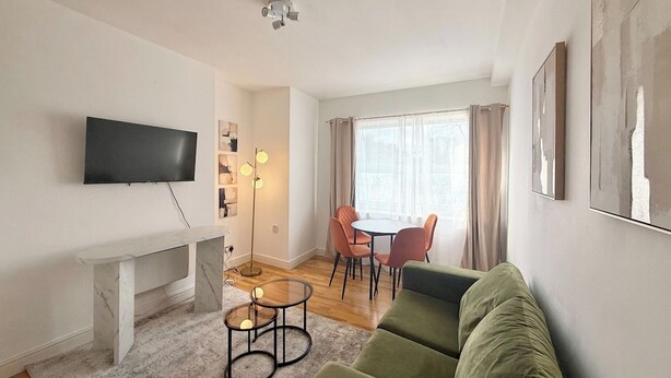 Una sala de estar moderna con un televisor de pantalla plana, una consola de mármol, un sofá verde, una mesa de centro de vidrio, una zona de comedor con mesa y sillas, y un ventanal con cortinas.