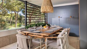 Comedor con mesa de madera preparada para cuatro, rodeada de sillas de mimbre, una lámpara colgante de fibra natural y vista a los árboles a través de un ventanal grande.
