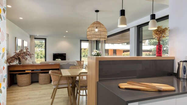 Una cocina moderna con una isla central, comedor con sillas de mimbre y ventanas amplias.